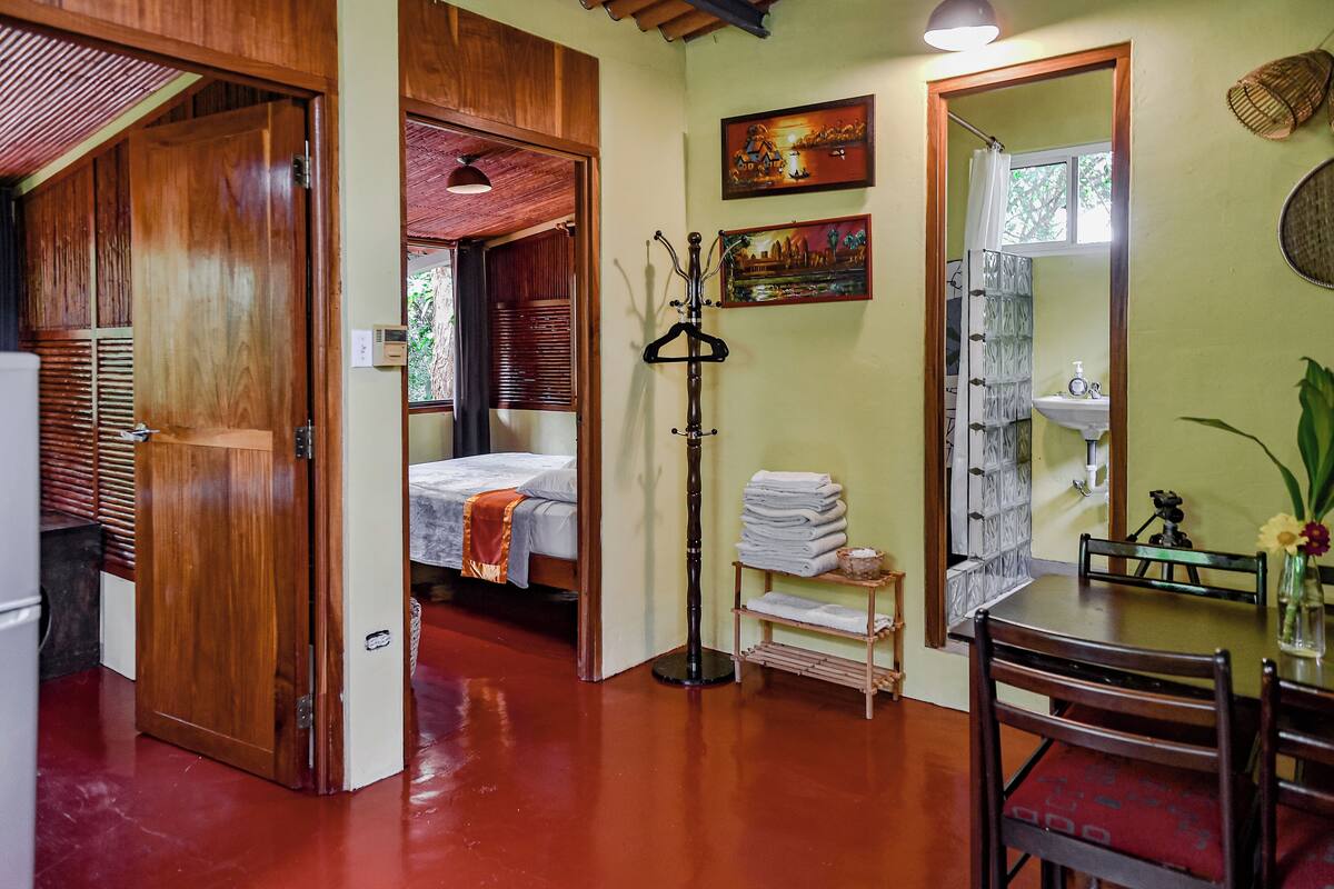 Una habitación con una cama, una puerta de madera, un espejo, un estante con toallas dobladas y una mesita con un florero.