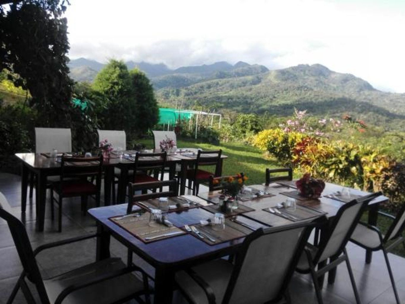 An outdoor dining area with tables and chairs, set against a backdrop of mountains and greenery.