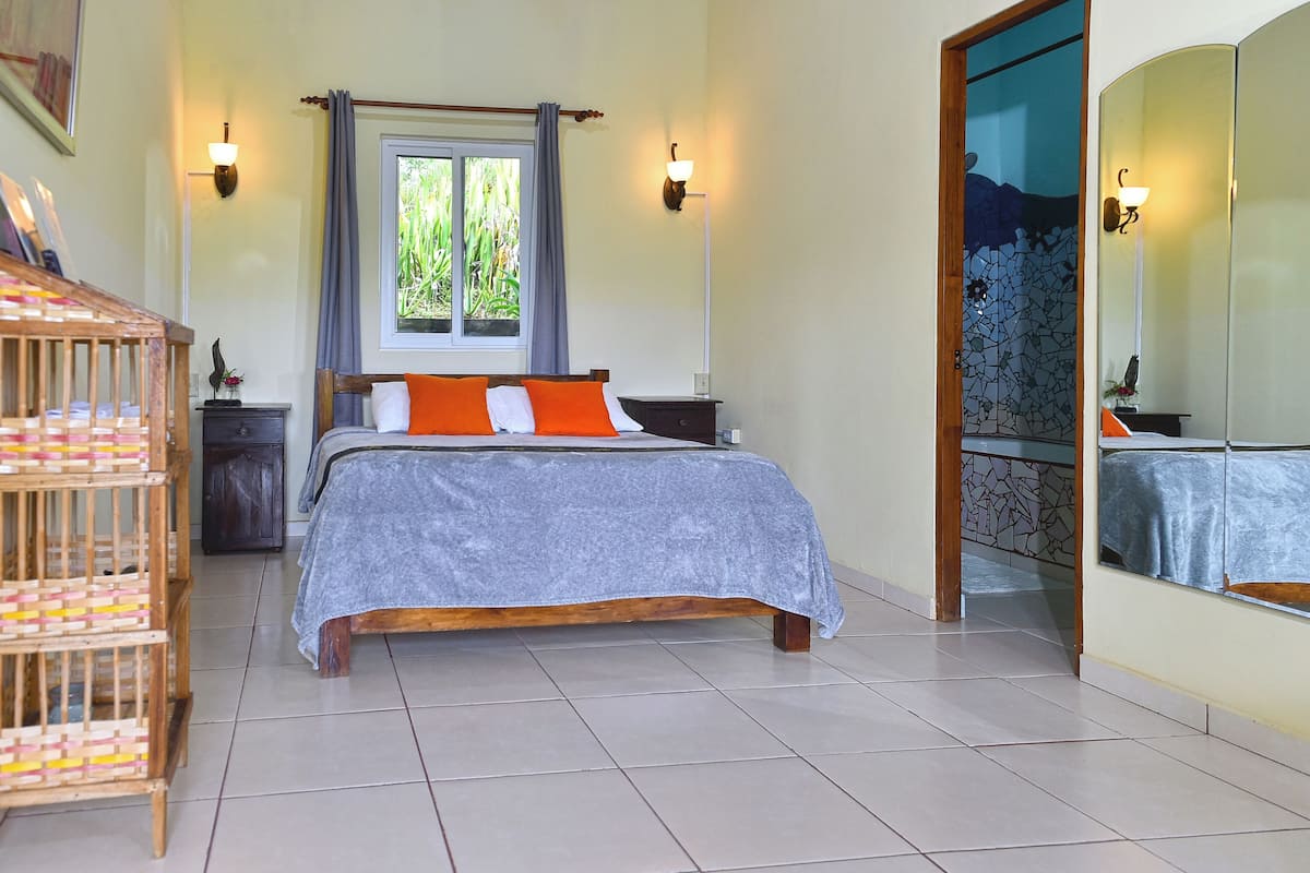 Un dormitorio con cama, mesita de noche, cómoda, ventana con cortinas, espejo y suelo de baldosas.