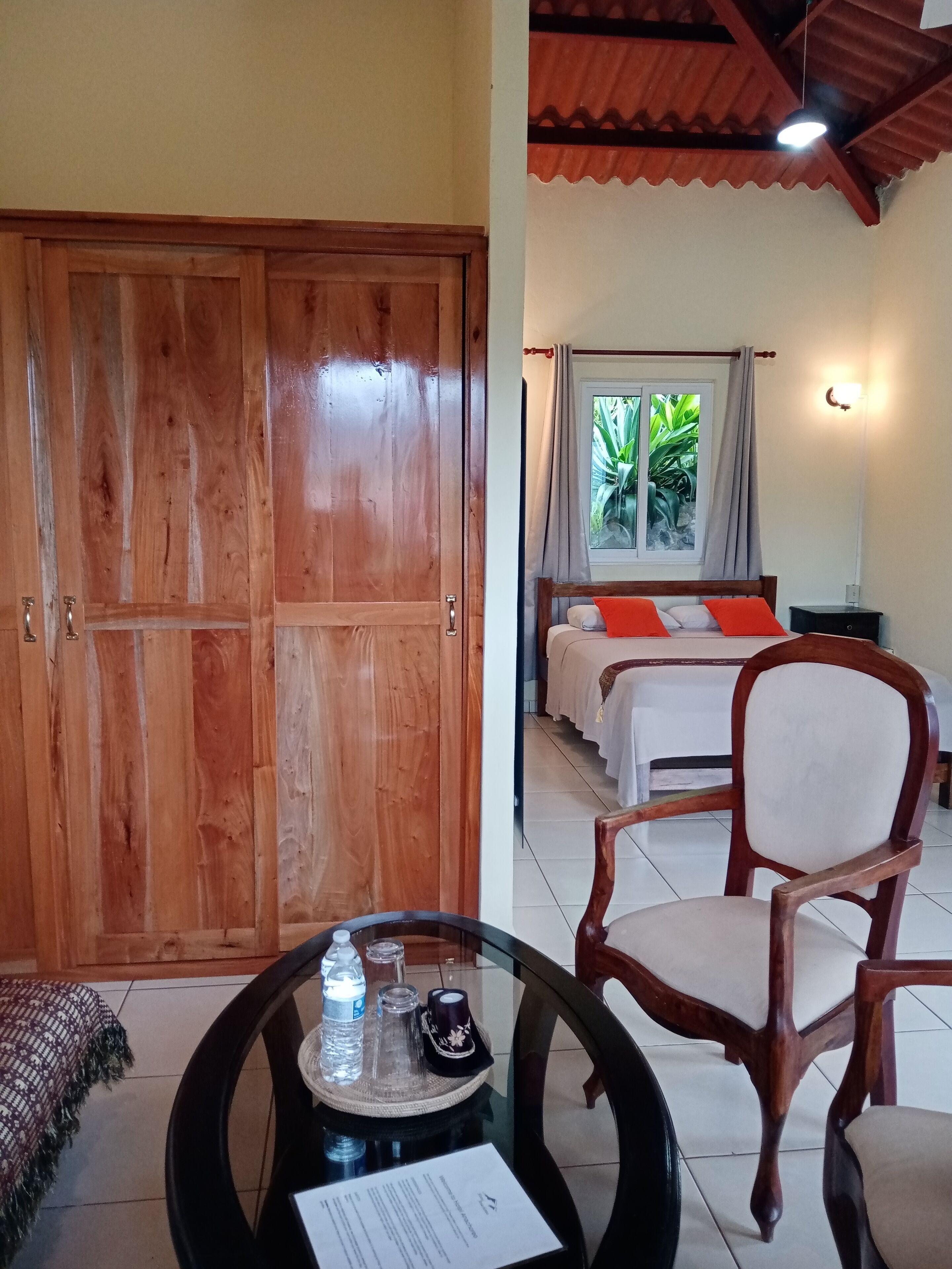 Quarto com guarda-roupa de madeira, cama com almofadas laranja, uma cadeira, mesa com uma garrafa de água e um menu, e janela com vista para o verde.
