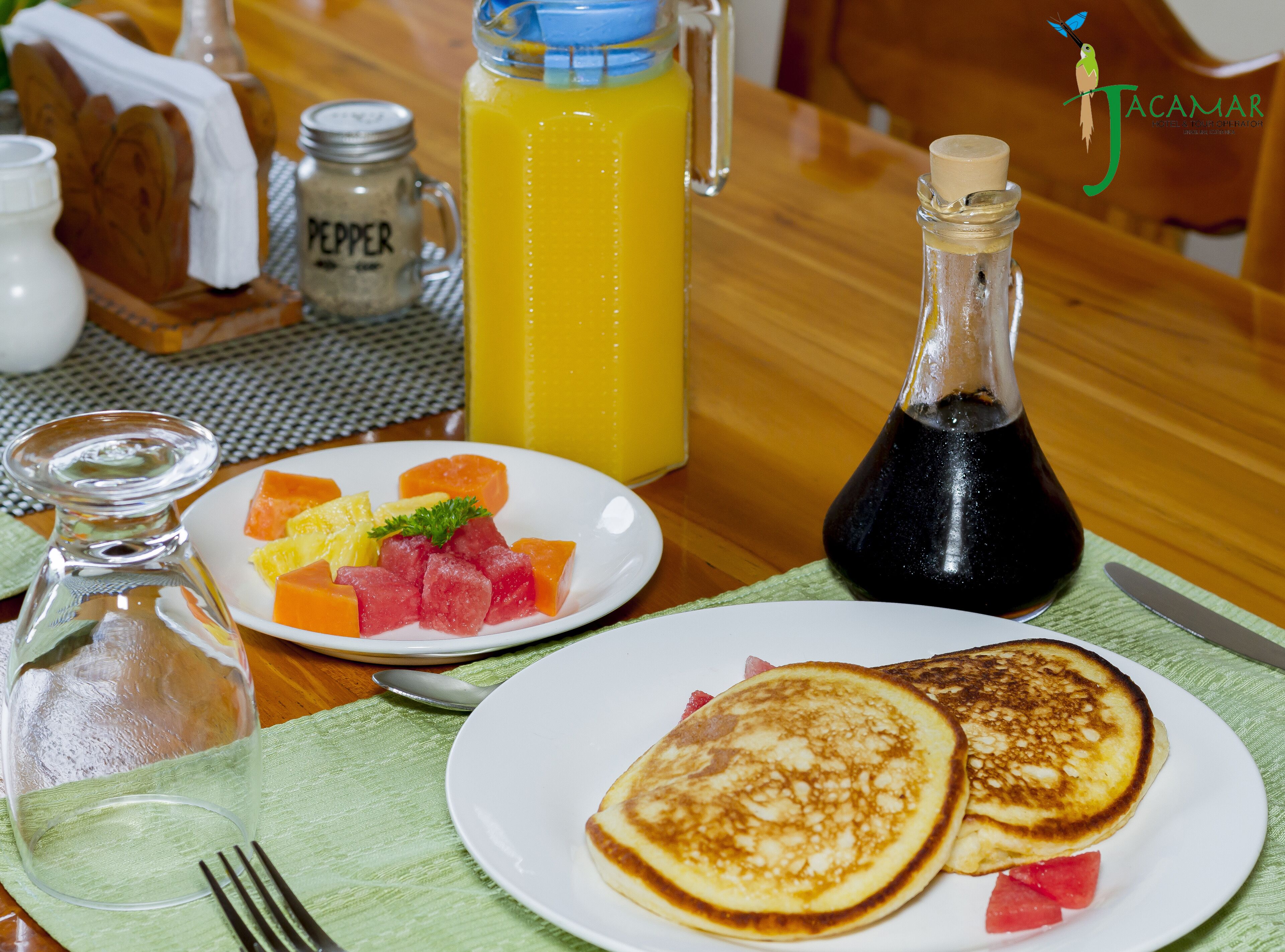 A breakfast setting with pancakes, fruit, and a bottle of syrup on a wooden table.