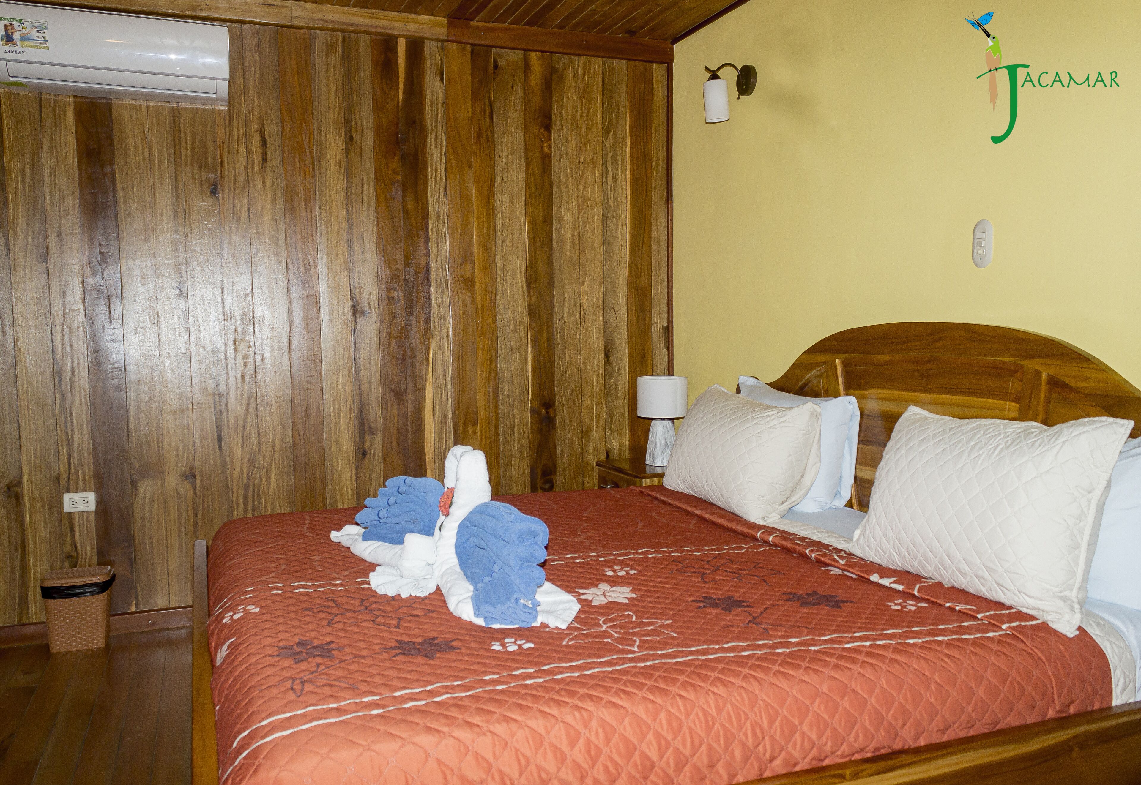 A hotel room with a wooden paneled wall, a bed with a red quilted bedspread, white pillows, and a wooden headboard.