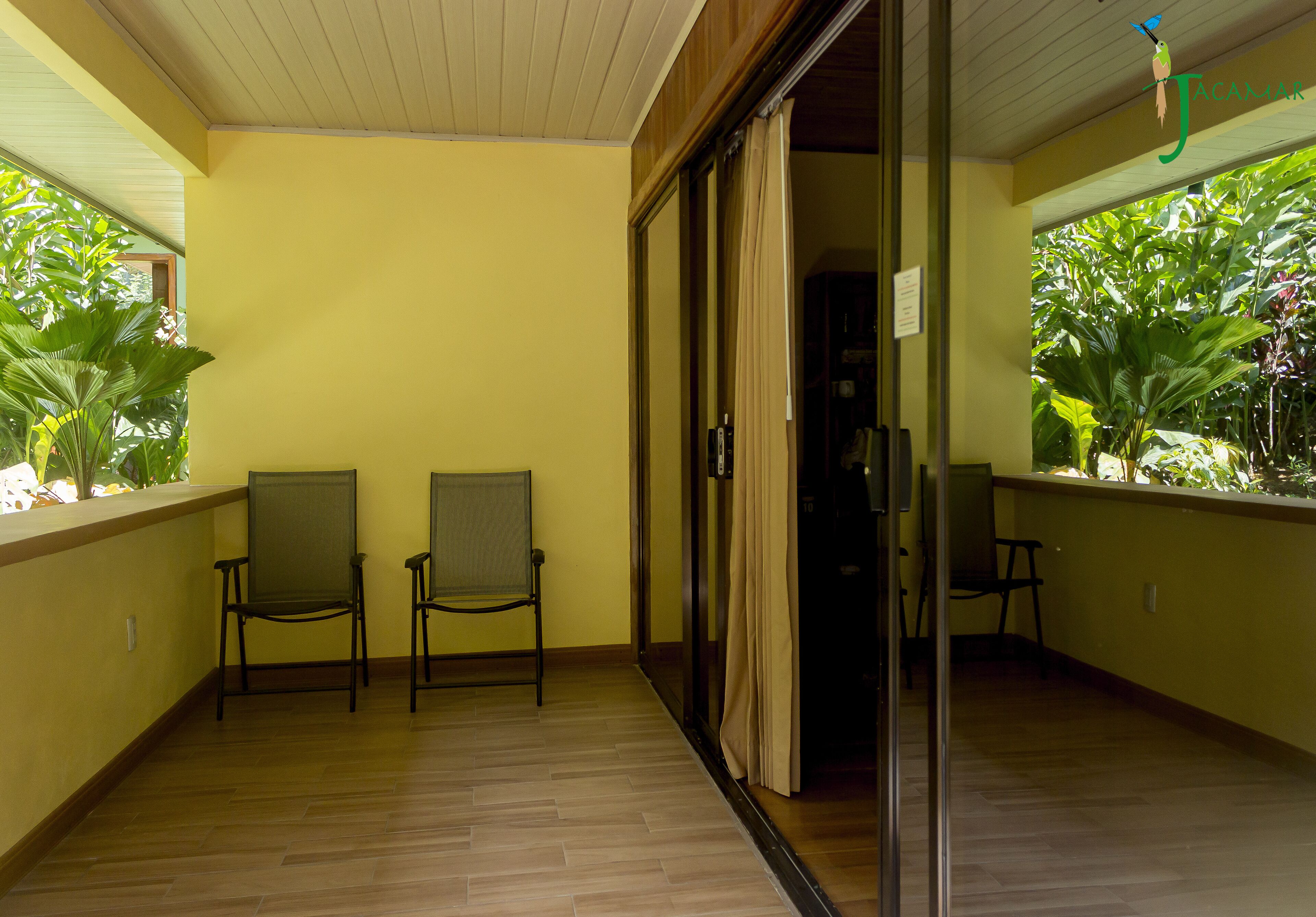 A balcony with two chairs, a sliding glass door, and a view of lush greenery.
