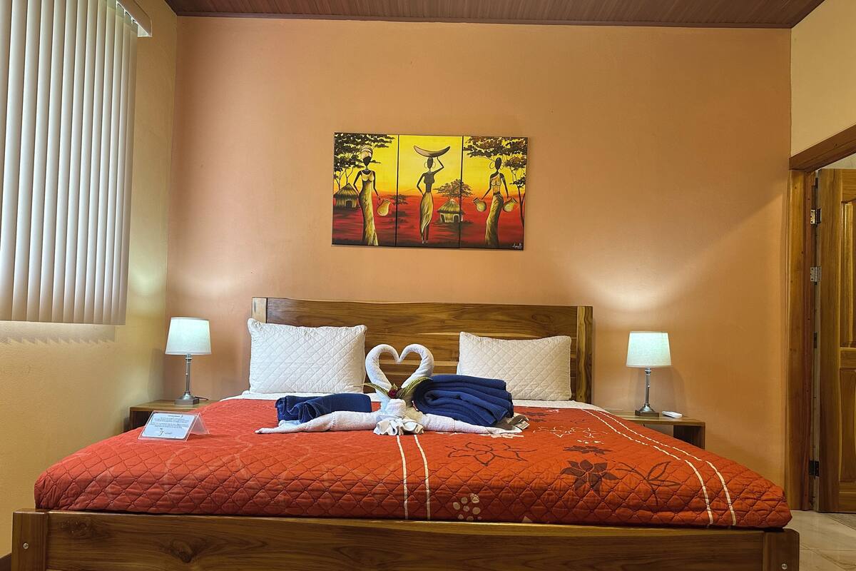 A neatly made bed with a red quilt, two white pillows, and a heart-shaped towel arrangement. There is a painting of women on the wall above the bed.