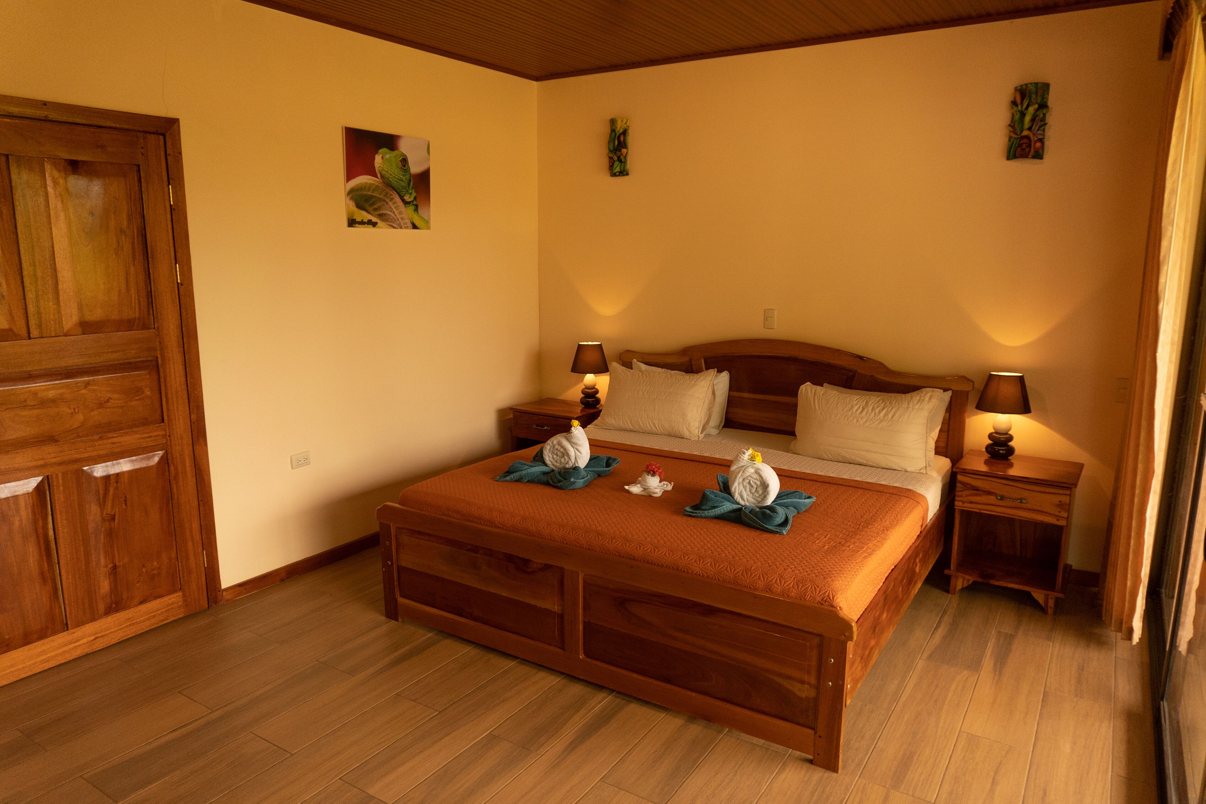 A bedroom with a wooden bed, two bedside tables with lamps, a wooden wardrobe, and two wall-mounted paintings.