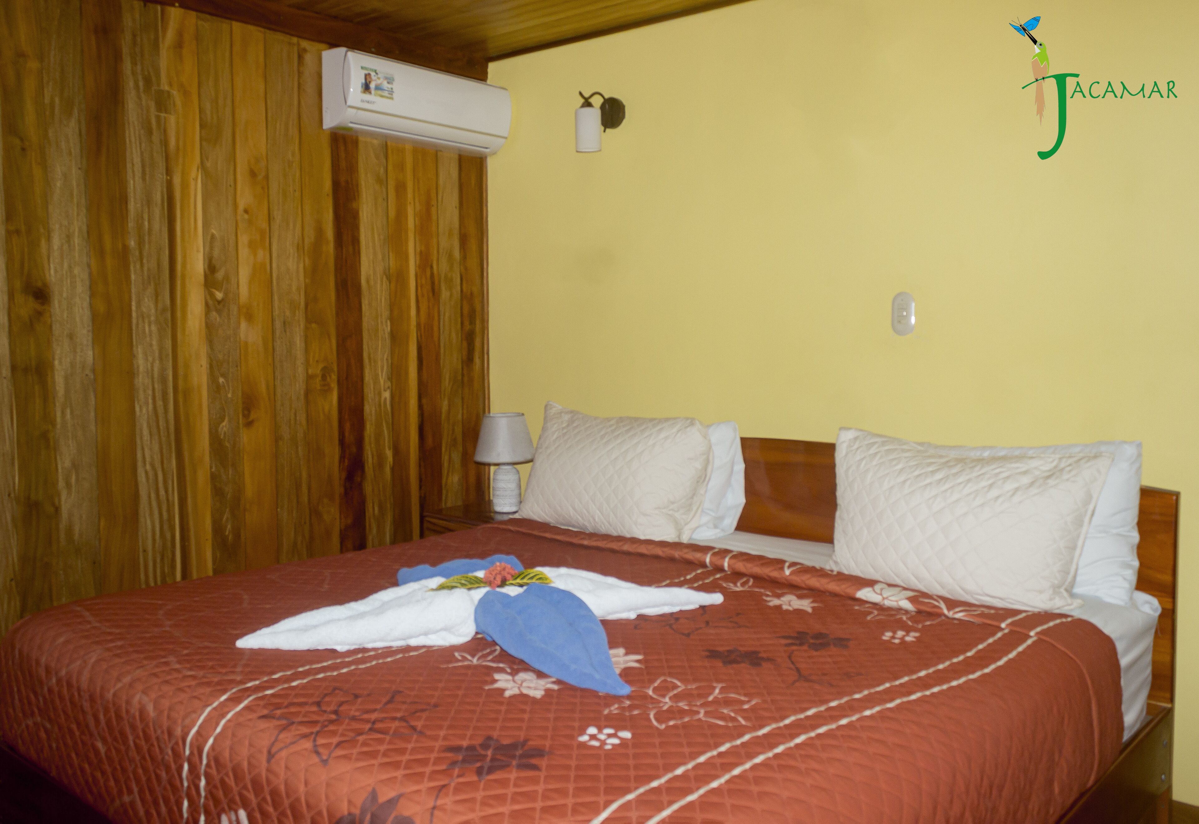 A hotel room with a wooden ceiling, a bed with a floral bedspread, white pillows, and a nightstand with a lamp.