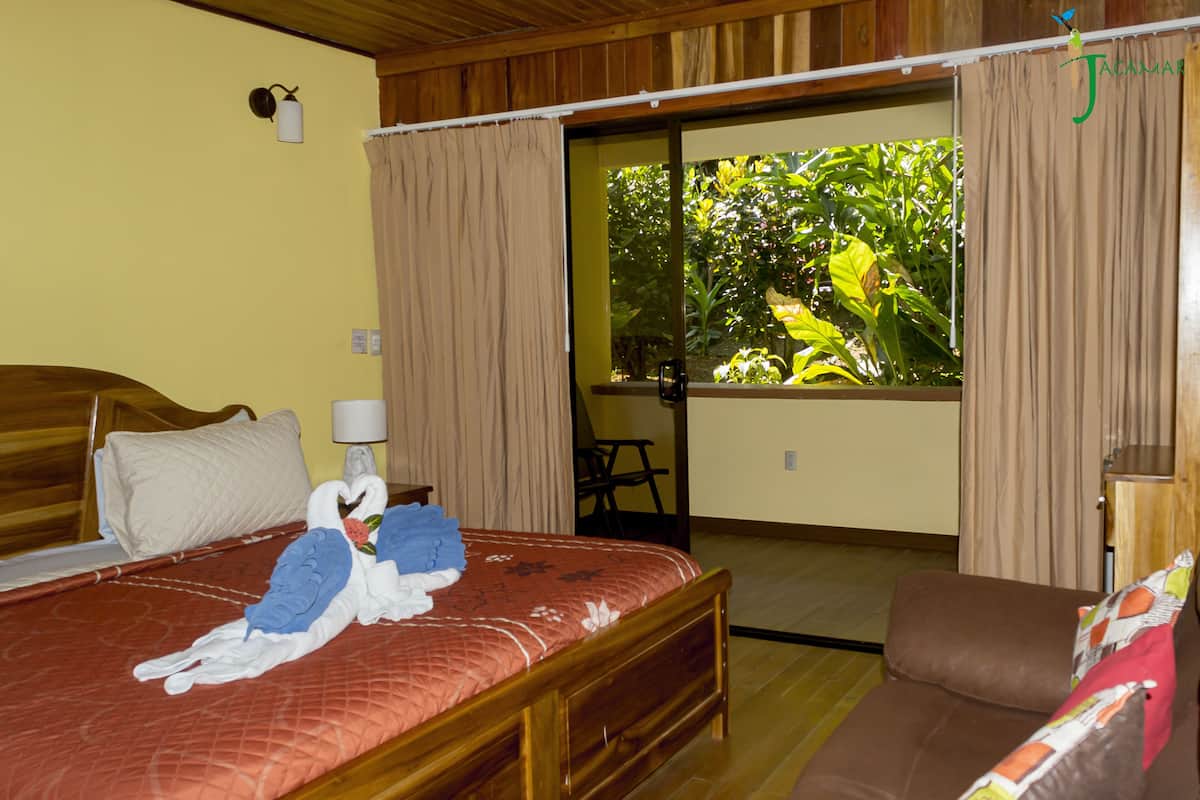 A hotel room with a bed, a sofa, a window with curtains, and a view of greenery outside.
