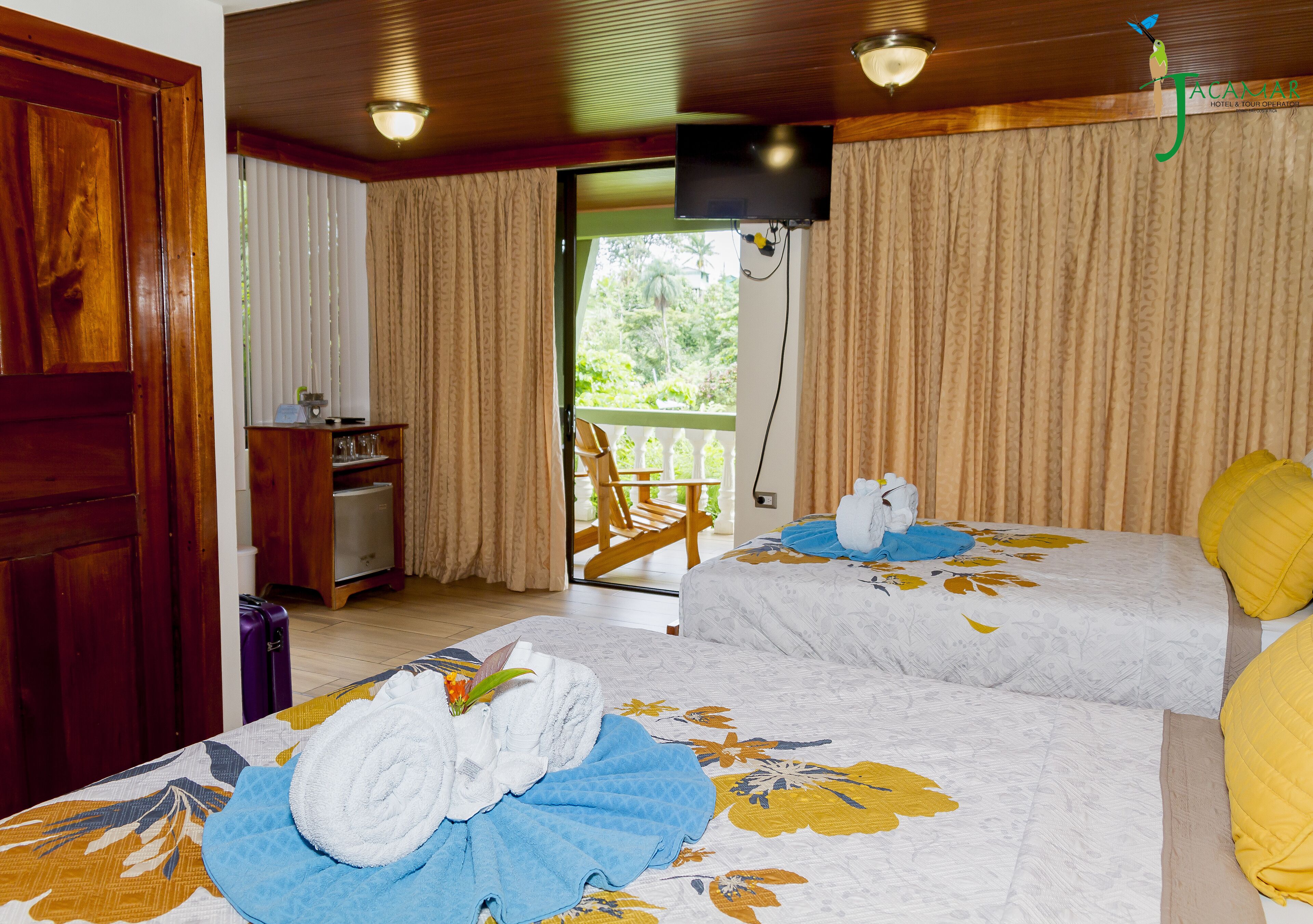 A hotel room with two beds, a TV, a desk with a computer, and a balcony with a view of greenery.