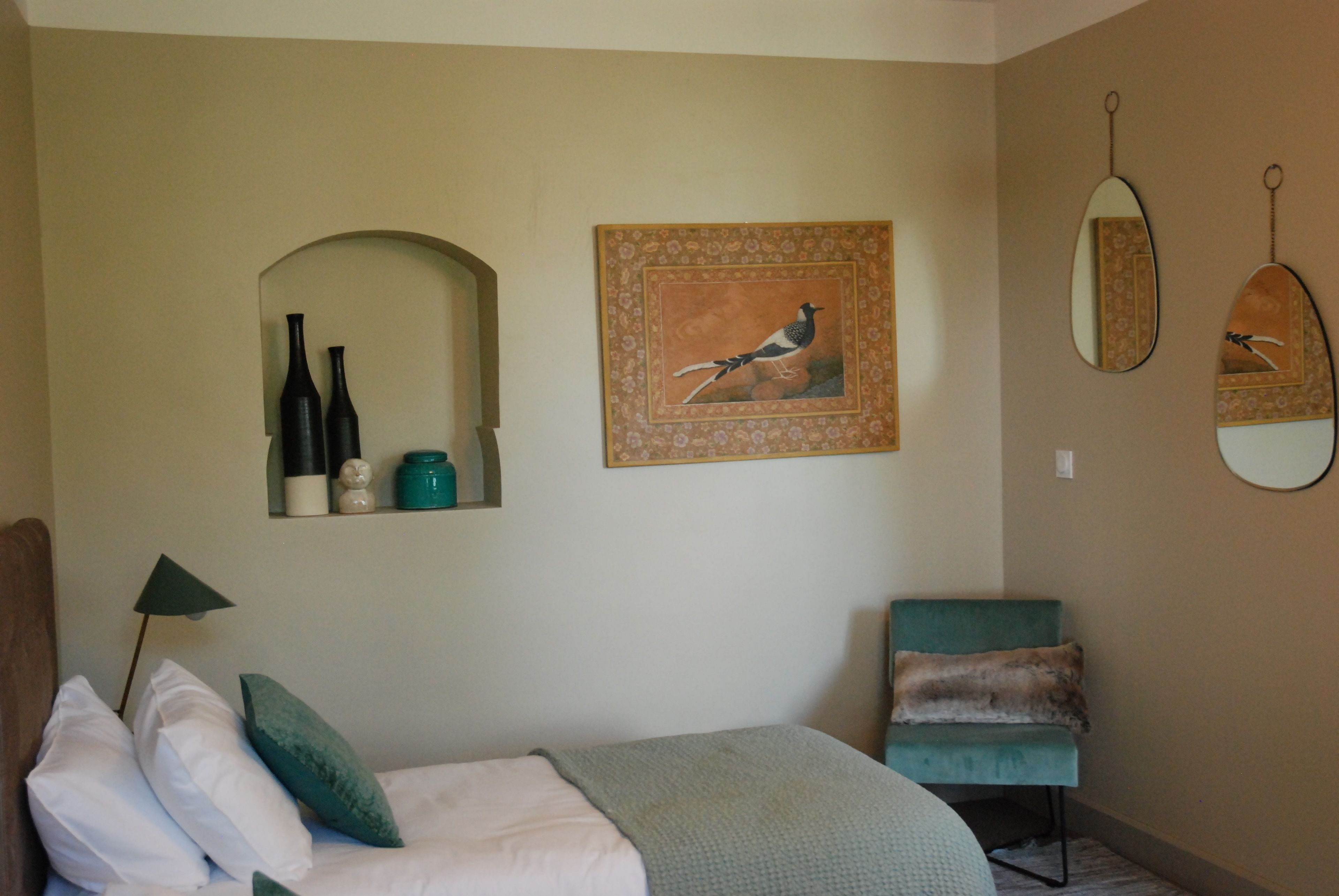 Een slaapkamer met een bed, kussens, een bedlampje, een stoel, een spiegel en wandplanken met flessen en een decoratief object.