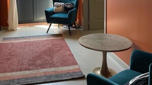 A room with a teal armchair, a round table, and a view through a window with sheer curtains.
