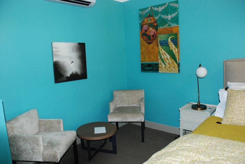 Una habitación con paredes turquesa, una cama con colcha amarilla y blanca, un sillón gris, una pequeña mesa redonda con un objeto blanco encima y una mesita de noche con una lámpara.
