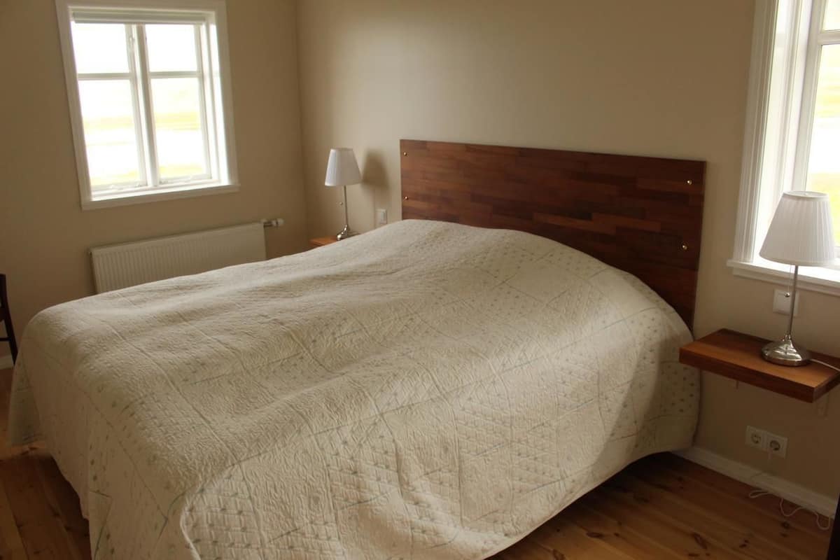 Un dormitorio con una cama grande, un cabecero de madera, una mesita de noche con una lámpara y una ventana que deja entrar luz natural.