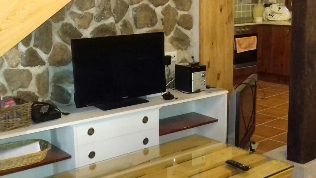 Una habitación con pared de piedra, un televisor de pantalla plana sobre un mueble blanco, una mesa de madera con un control remoto y una zona de cocina al fondo.