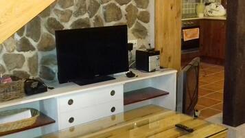 Una habitación con pared de piedra, un televisor de pantalla plana sobre un mueble blanco, una mesa de madera con un control remoto y una zona de cocina al fondo.