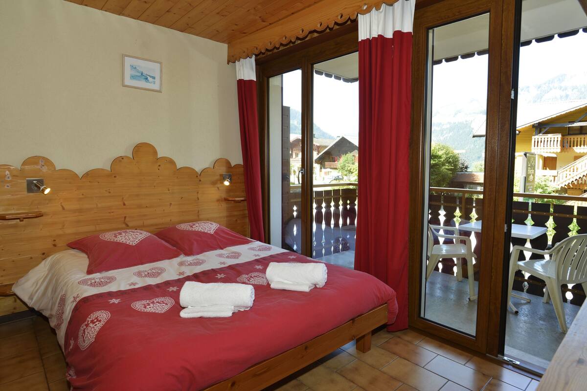 Une chambre d’hôtel avec une tête de lit en bois, un grand lit avec une literie rouge et un balcon aménagé avec du mobilier d’extérieur.