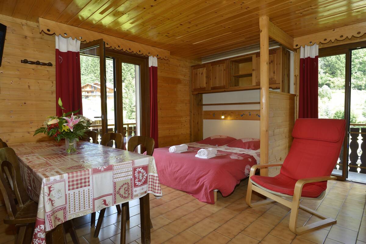 Une pièce de cabane en bois comprenant une table à manger, un lit avec une literie rouge, une chaise rouge et une fenêtre avec des rideaux.