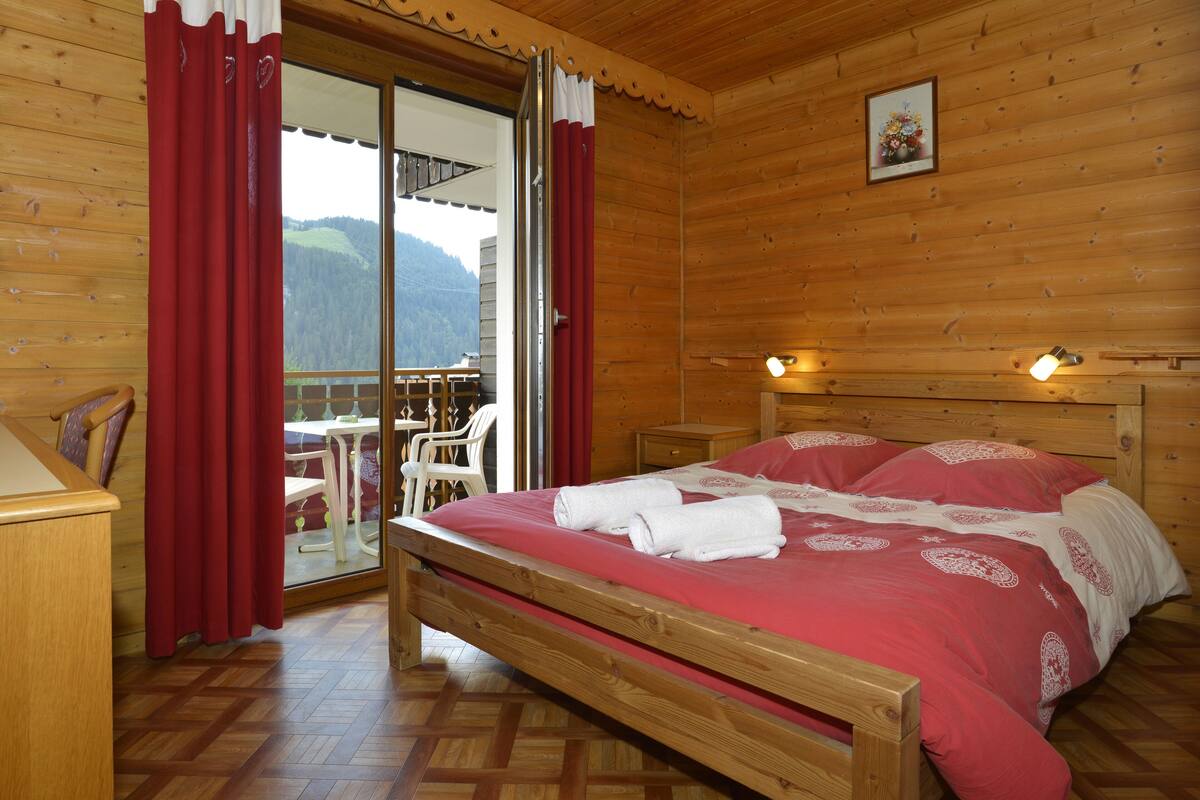 Une chambre à coucher dans une cabane en bois, avec un lit, des rideaux rouges, une tête de lit en bois et une vue sur l’extérieur.