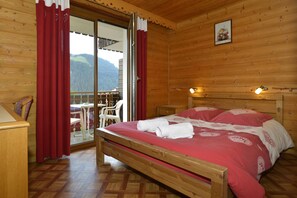 A wooden cabin bedroom with a bed, red curtains, a wooden headboard, and a view of the outdoors. - La Fleche D'or (Chatel)