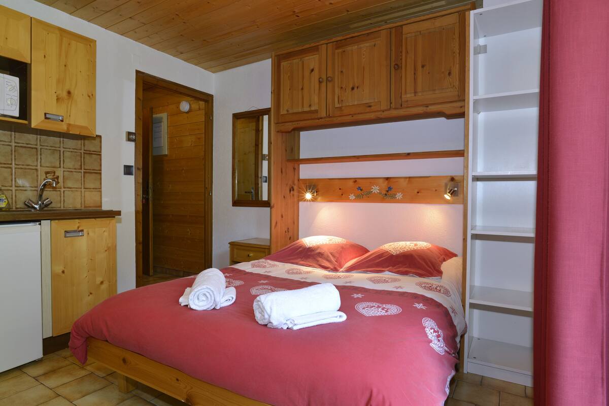 Une chambre avec un lit en bois, une table de chevet intégrée, une armoire en bois et un petit coin cuisine.