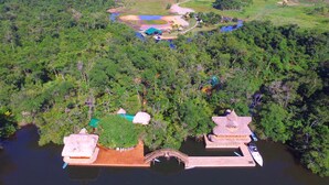 An aerial view of a floating structure with two thatched-roof buildings connected by a wooden bridge over water, surrounded by dense forest. - Tenamit Maya (Livingston)