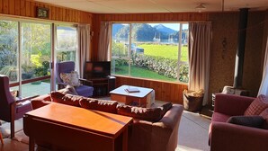 A cozy living room with a wooden interior, a television, a sofa, and a table. Large windows offer a view of a green landscape and a mountain. - Ngakuta Bay, Marlborough Sounds. (Ngakuta Bay)