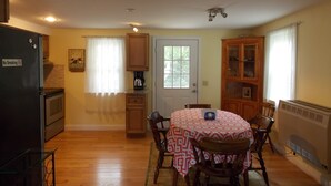 Un comedor con una mesa cubierta con un mantel estampado, rodeada de sillas de madera. Se ve un refrigerador a la izquierda y un armario con puertas de vidrio a la derecha.