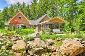 A cabin with a covered patio, surrounded by a natural landscape with large rocks and dense trees. - Hot Tub, Dock, View! Waterfront Great Pond Cottage (Great Pond)