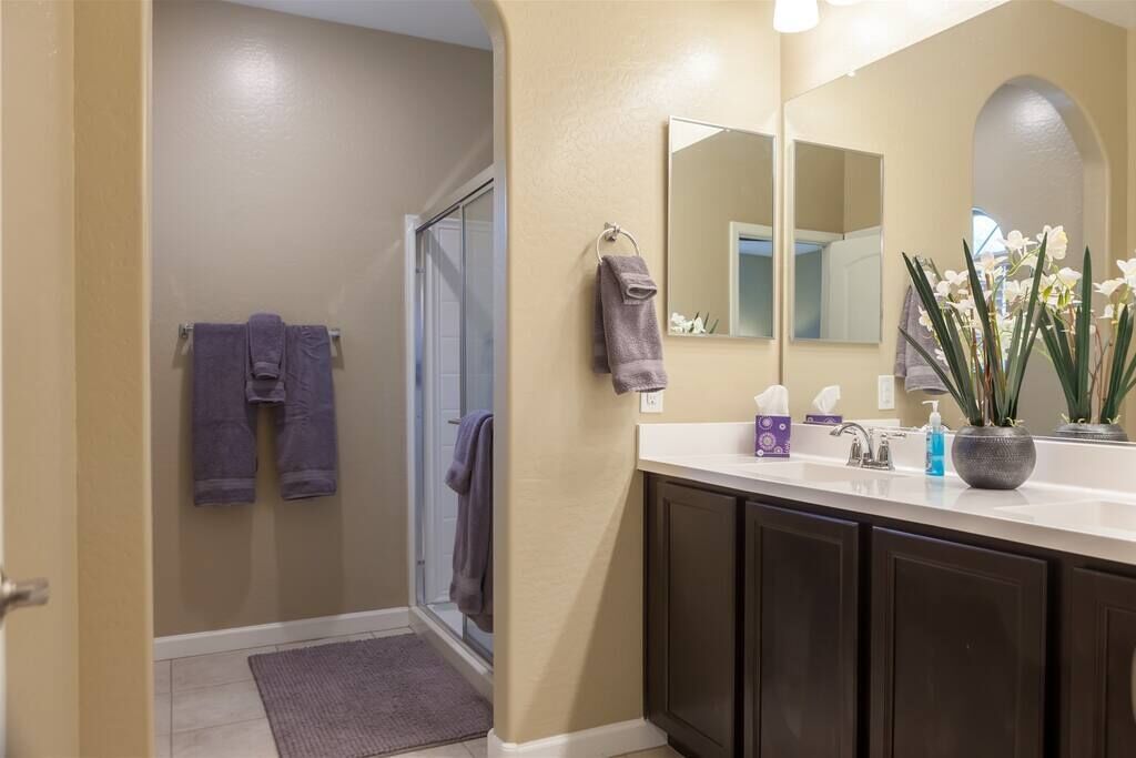 A bathroom with a large mirror, dark cabinets, a sink with toiletries, and a door leading to a shower area.