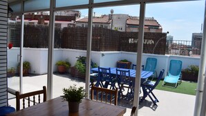 A patio area with a wooden table and blue chairs, a small potted plant, and a view of neighboring buildings and a fence. - Duplex of 90m2, 6 persons with terrace of 30m2 (Barcelona)