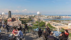 An outdoor dining area with tables and chairs, overlooking a cityscape with buildings and a body of water. - Duplex of 90m2, 6 persons with terrace of 30m2 (Barcelona)