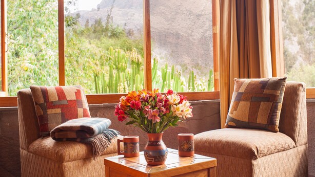 A cozy seating area with two cushioned chairs, a small table with a vase of flowers, and a view of greenery through large windows. - Willka T'ika Essential Wellness (Urubamba)