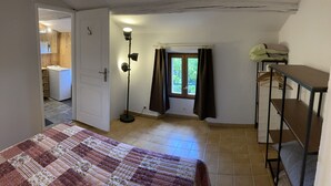 Een slaapkamer met een bed, nachtkastje, vloerlamp, raam met gordijnen en uitzicht op een keuken en badkamer.