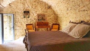 Un dormitorio con pared de piedra, una cama, un escritorio de madera, dos sillas y un cuadro en la pared.