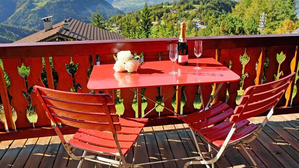 Une table rouge avec deux chaises sur un balcon donnant sur un paysage montagneux.