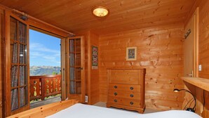 Une chambre dans une cabane en bois, avec un lit, une commode et une vue sur les montagnes par une porte ouverte.