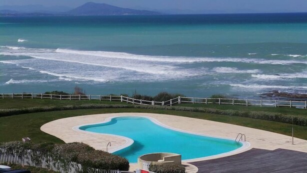 A swimming pool with a curved design, surrounded by a wooden deck and a white fence. - Luxury studio 1st line Ocean for 2 persons (Biarritz)