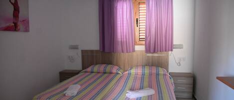 A room with two beds, a wooden headboard, a window with purple curtains, a bedside table, and a wall-mounted painting.