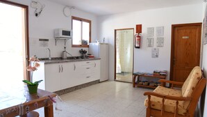 A kitchen with white cabinets, a microwave, and a refrigerator. There is a dining table with chairs and a potted plant. - Pensión Eva (Mogan)