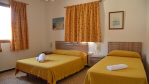 A hotel room with two beds, each with a yellow bedspread and a rolled towel. There is a painting on the wall and a ceiling fan. - Pensión Eva (Mogan)