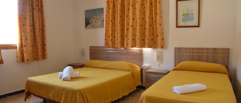 A hotel room with two beds, each with a yellow bedspread and a rolled towel. There is a painting on the wall and a ceiling fan.