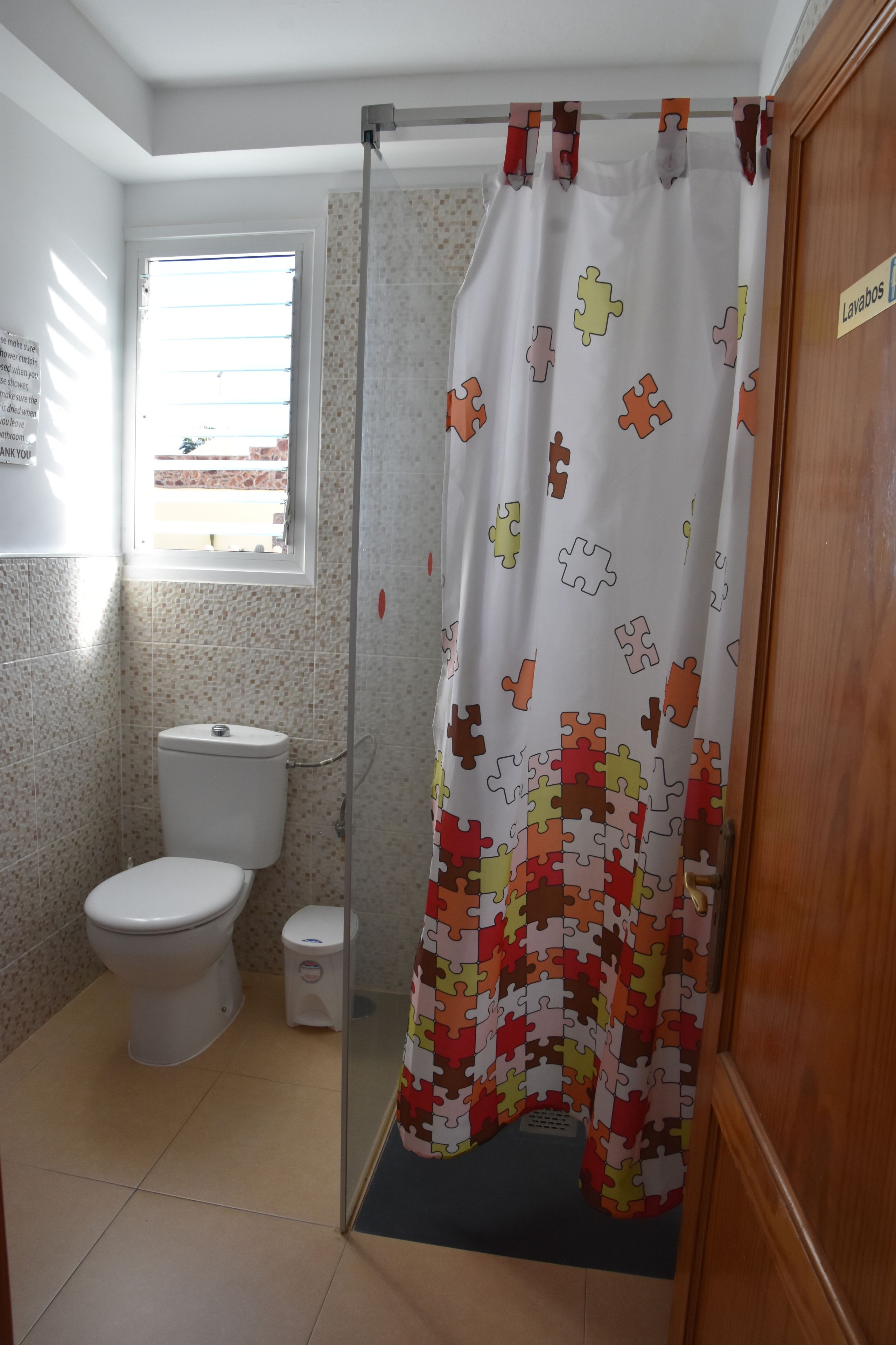 A bathroom with a toilet, a small trash bin, a shower curtain with a puzzle pattern, and a window with blinds.