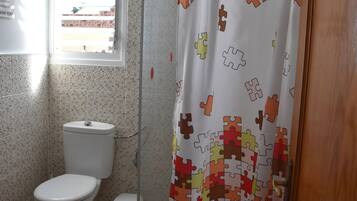 A bathroom with a toilet, a small trash bin, a shower curtain with a puzzle pattern, and a window with blinds.