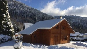 Uma cabana de madeira com telhado coberto de neve, rodeada por árvores carregadas de neve e uma montanha arborizada ao fundo.