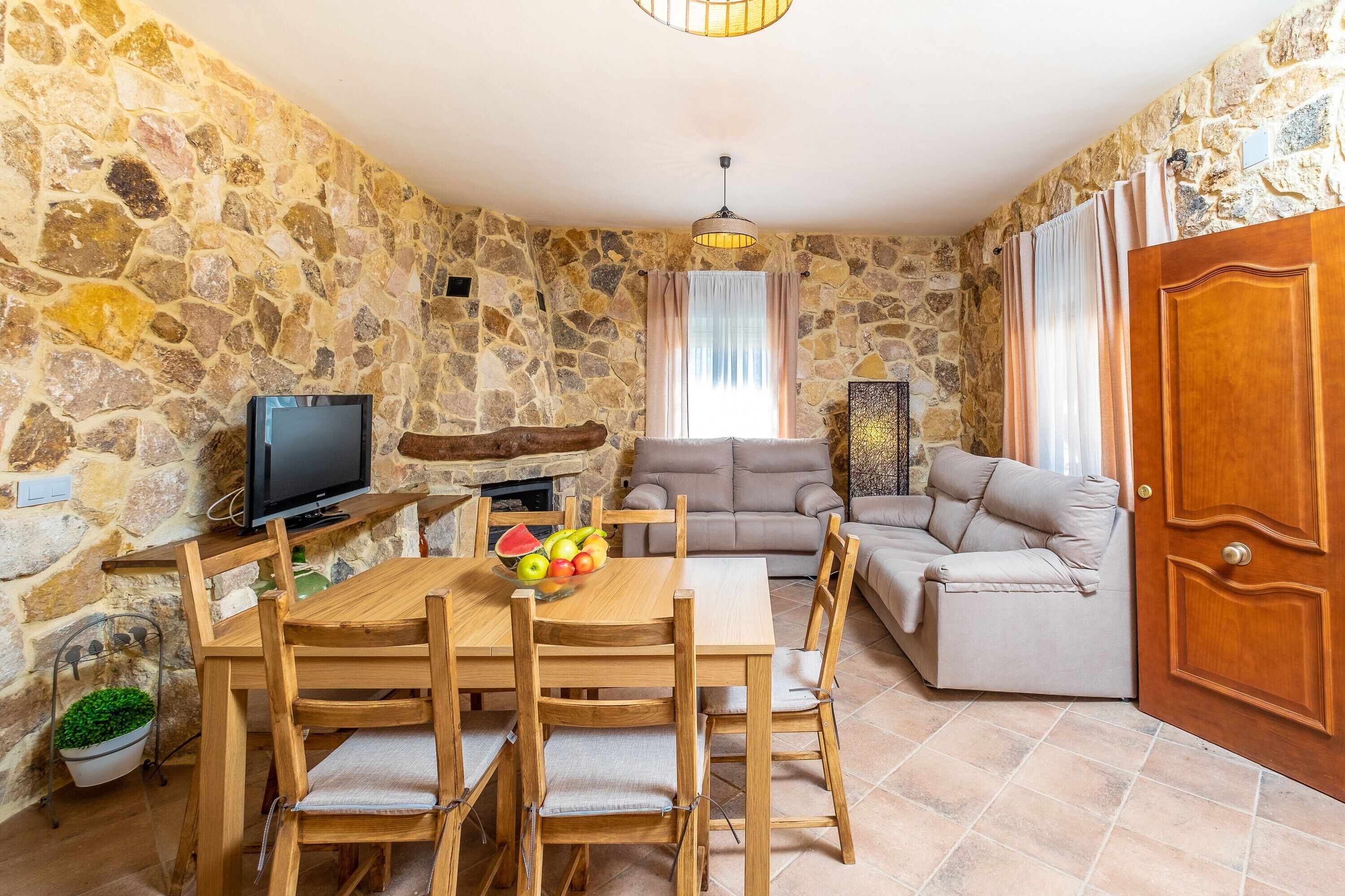 A stone-walled room with a wooden dining table, chairs, a television, a sofa, and a wooden door.