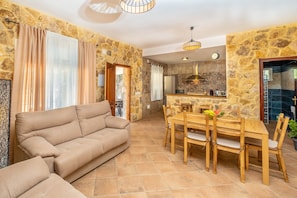Ein Wohnzimmer mit einer Couch, einem Esstisch mit Stühlen, einer Küchenzeile mit Kühlschrank und einer Steinwand.