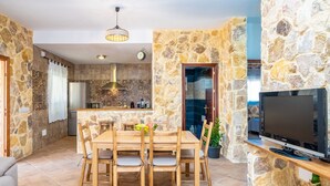 Un coin repas avec un mur en pierre, une cuisine à l’arrière-plan et un téléviseur posé sur un meuble en bois.