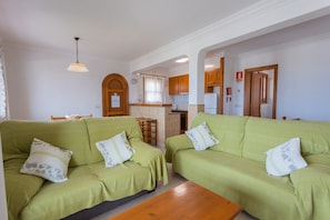 A living room with two green sofas, a wooden coffee table, and a kitchen in the background.