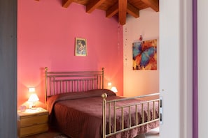 Quarto com cama, luminárias de cabeceira, mesa de cabeceira, pintura na parede e teto com vigas de madeira.