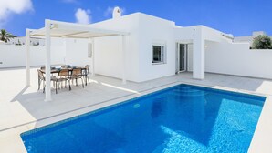 Ein modernes weißes Gebäude mit Swimmingpool, einer überdachten Terrasse mit Tisch und Stühlen und blauem Himmel.