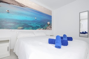 A hotel room with a large bed, white bedding, and a painting of a coastal scene.